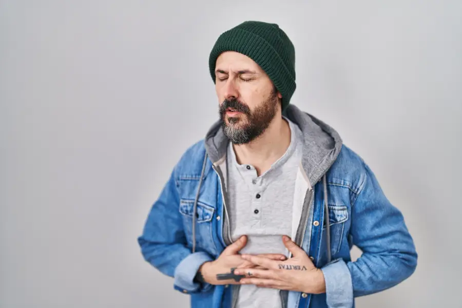 Young hispanic man with tattoos wearing wool cap with hand on stomach because indigestion painful illness feeling unwell ache concept